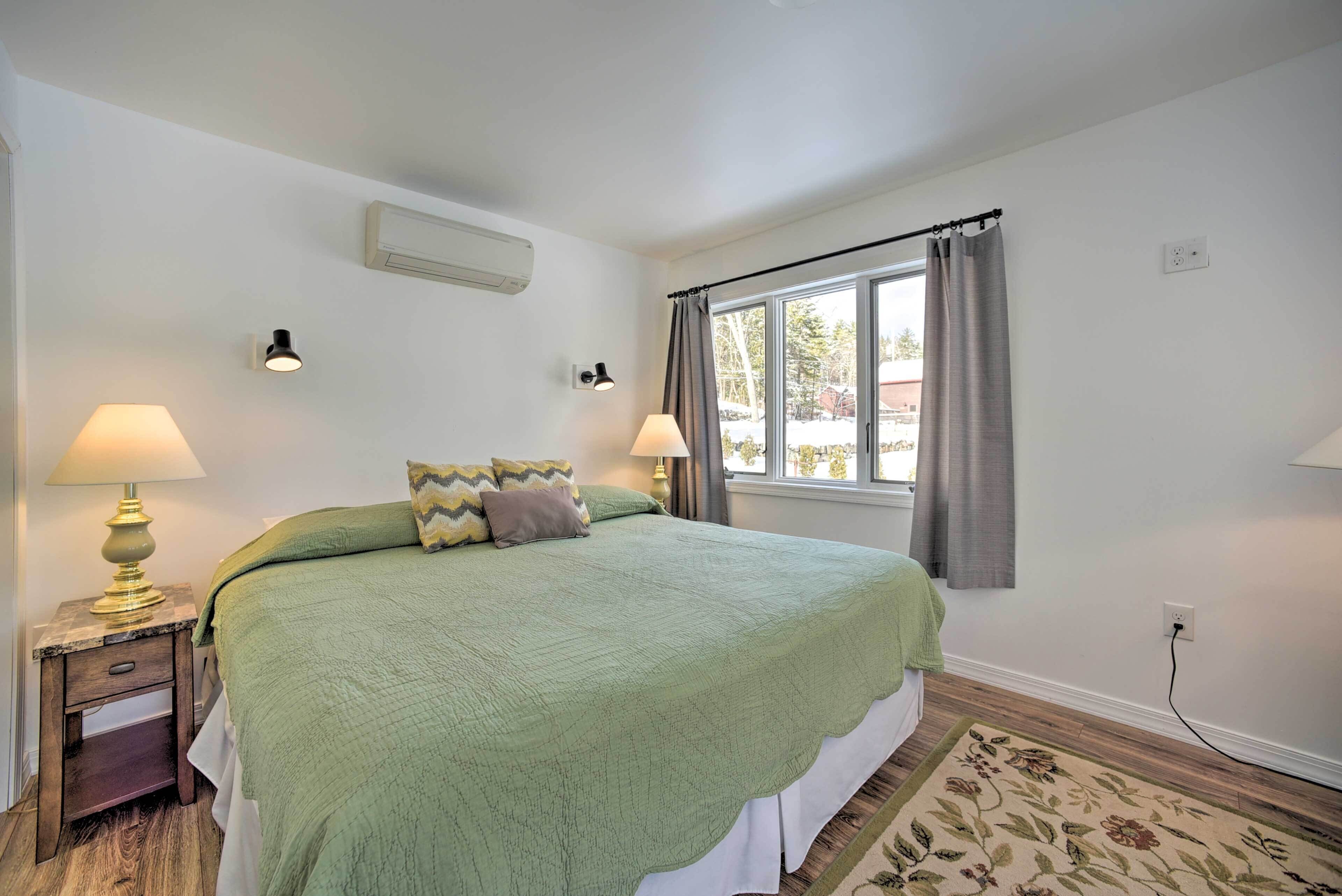 A bedroom with a large bed, two bedside lamps, a window with curtains, and a wall-mounted air conditioner.