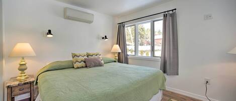 A bedroom with a large bed, two bedside lamps, a window with curtains, and a wall-mounted air conditioner.