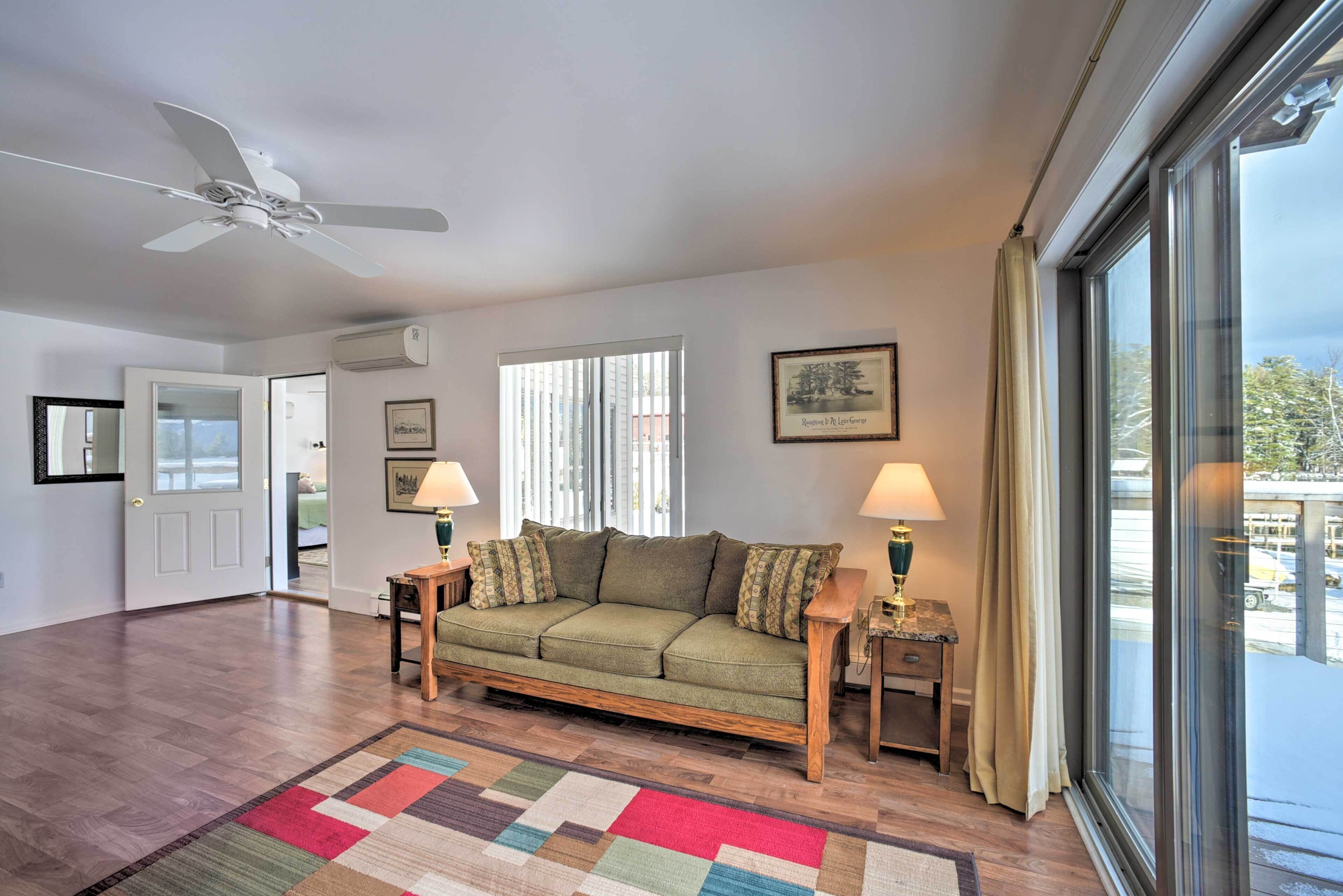 A spacious living room with a green sofa, wooden side tables, a ceiling fan, and large windows with curtains.