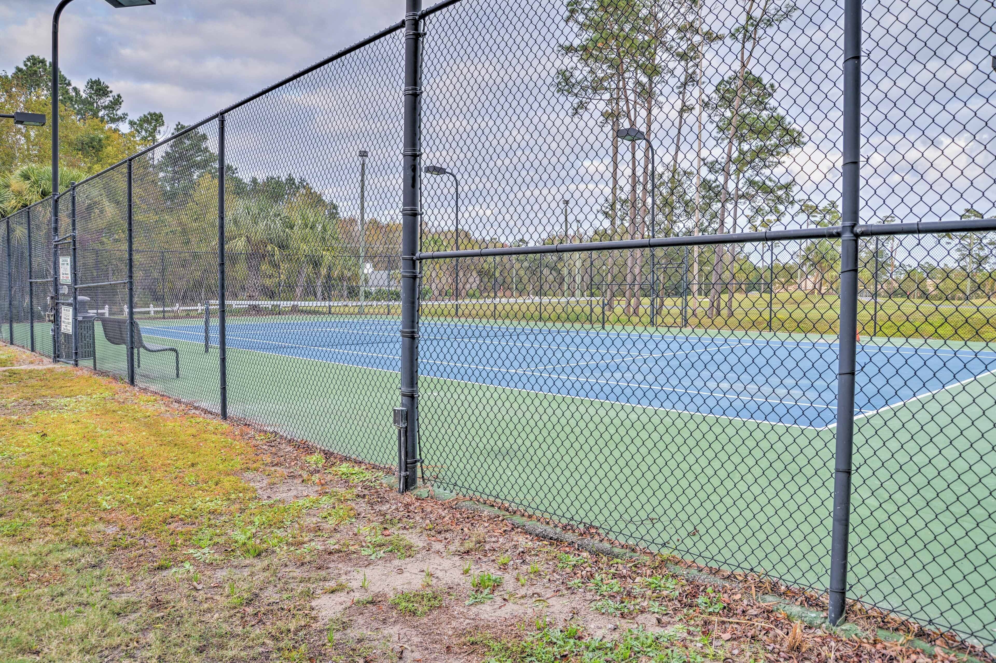 A tennis court enclosed by a chain-link fence with a green surface and a blue area, surrounded by trees and a cloudy sky.