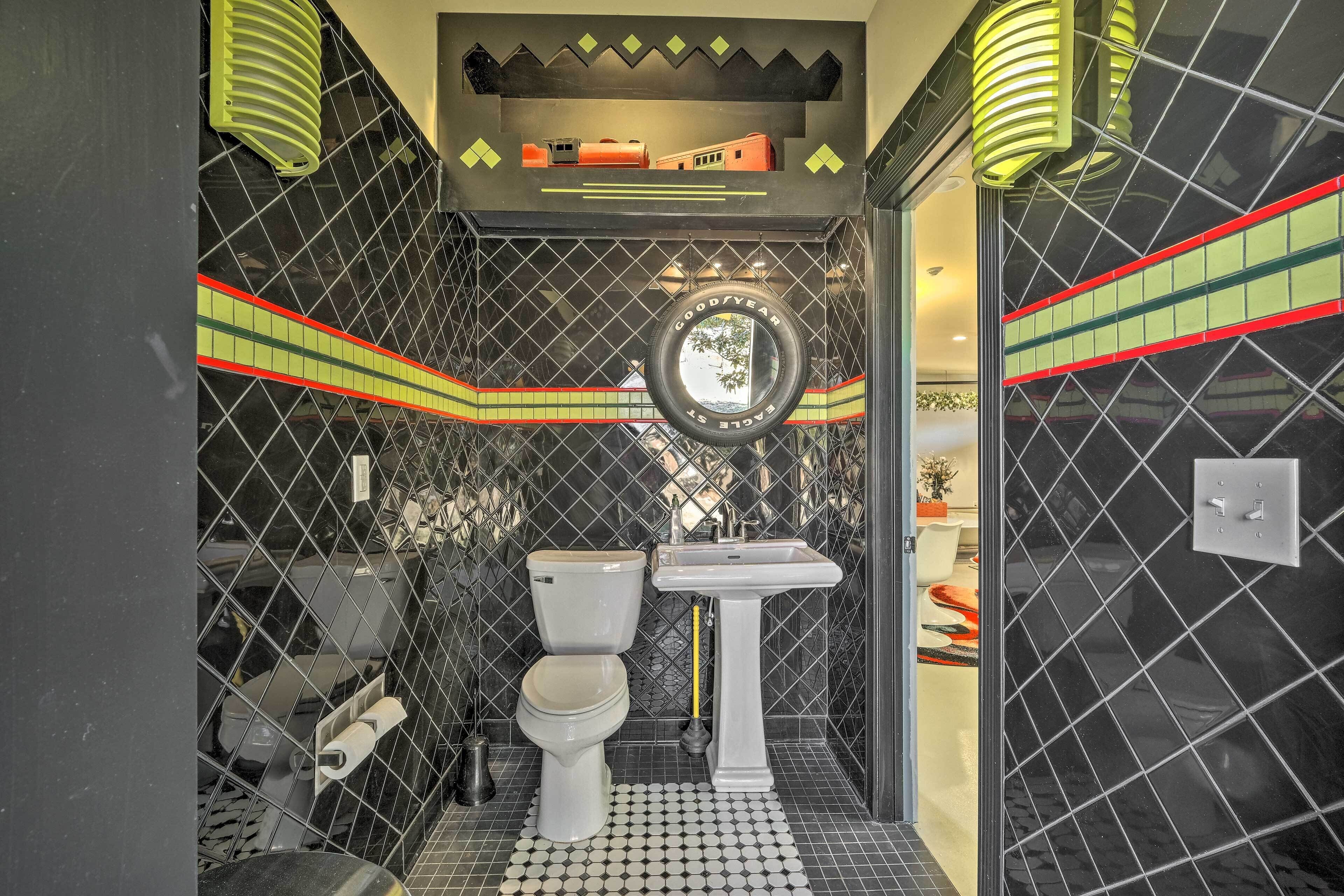 Un baño con pared de azulejos negros, un inodoro blanco, un lavabo y un espejo.