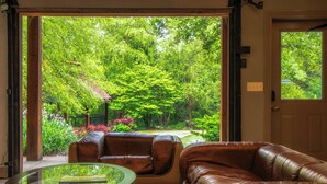 Un salon doté d’une table basse en verre, d’un canapé en cuir et de fauteuils, offrant une vue sur un jardin luxuriant et des arbres, le tout visible à travers une porte ouverte.