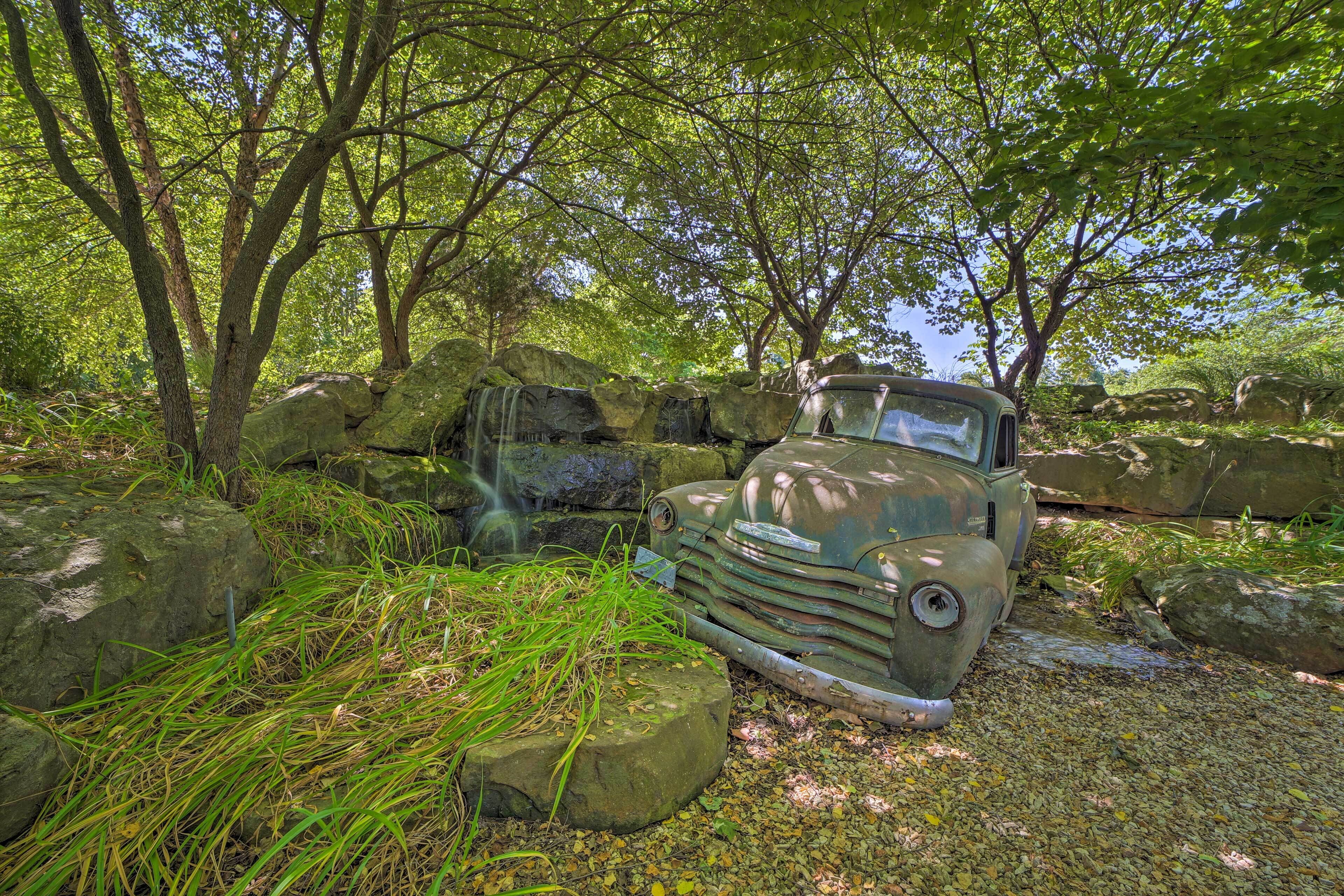 An old, abandoned car in a wooded area with large rocks and trees.