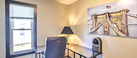 Een hotelkamer met een bureau, stoel, lamp, raam, prullenbak en een schilderij van een brug.