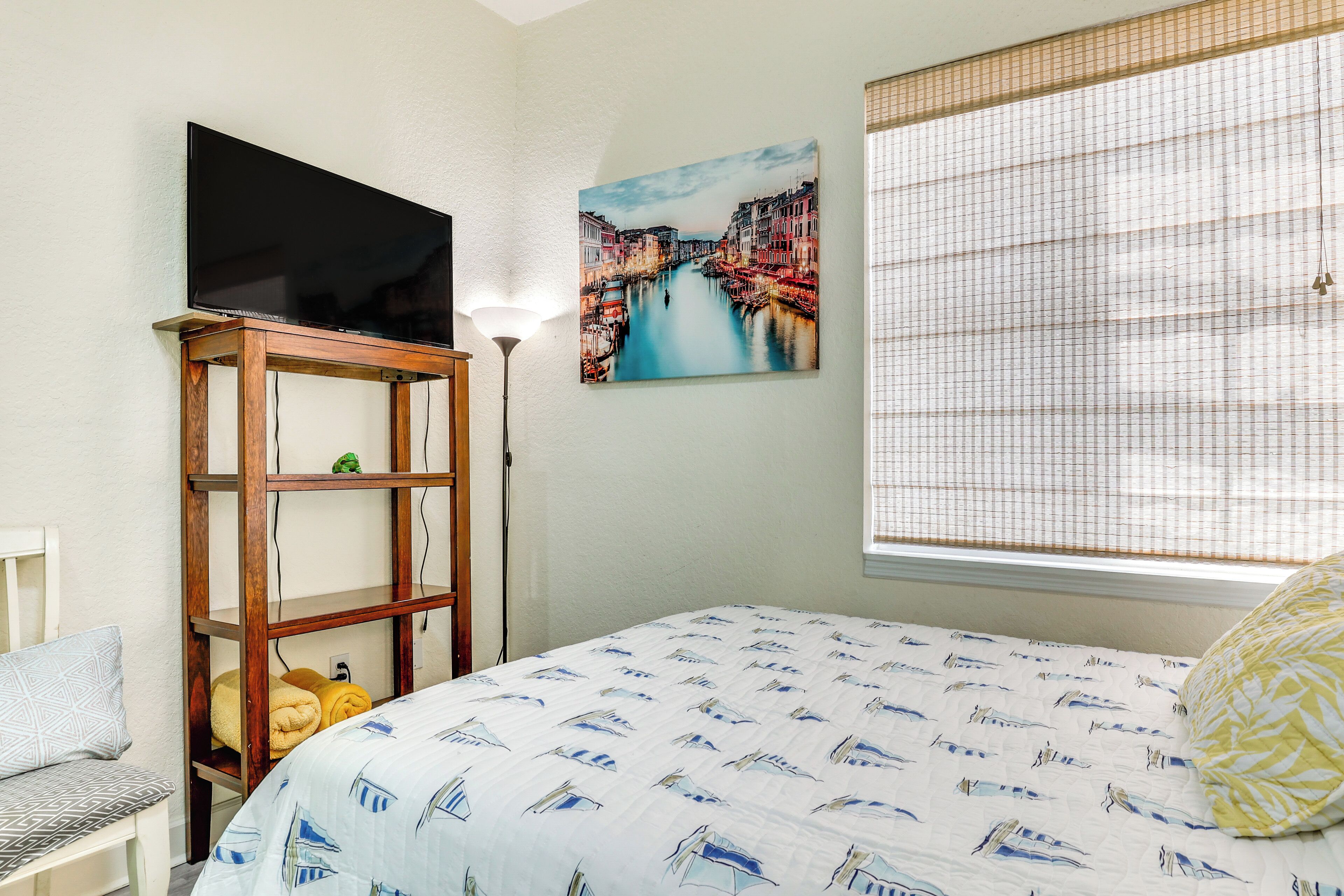 A bedroom with a bed, a TV on a wooden stand, a painting of a canal, and a window with blinds.