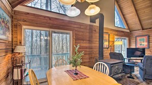 A cozy cabin interior with a wooden dining table, chairs, a fireplace, a TV, and a large window overlooking trees.