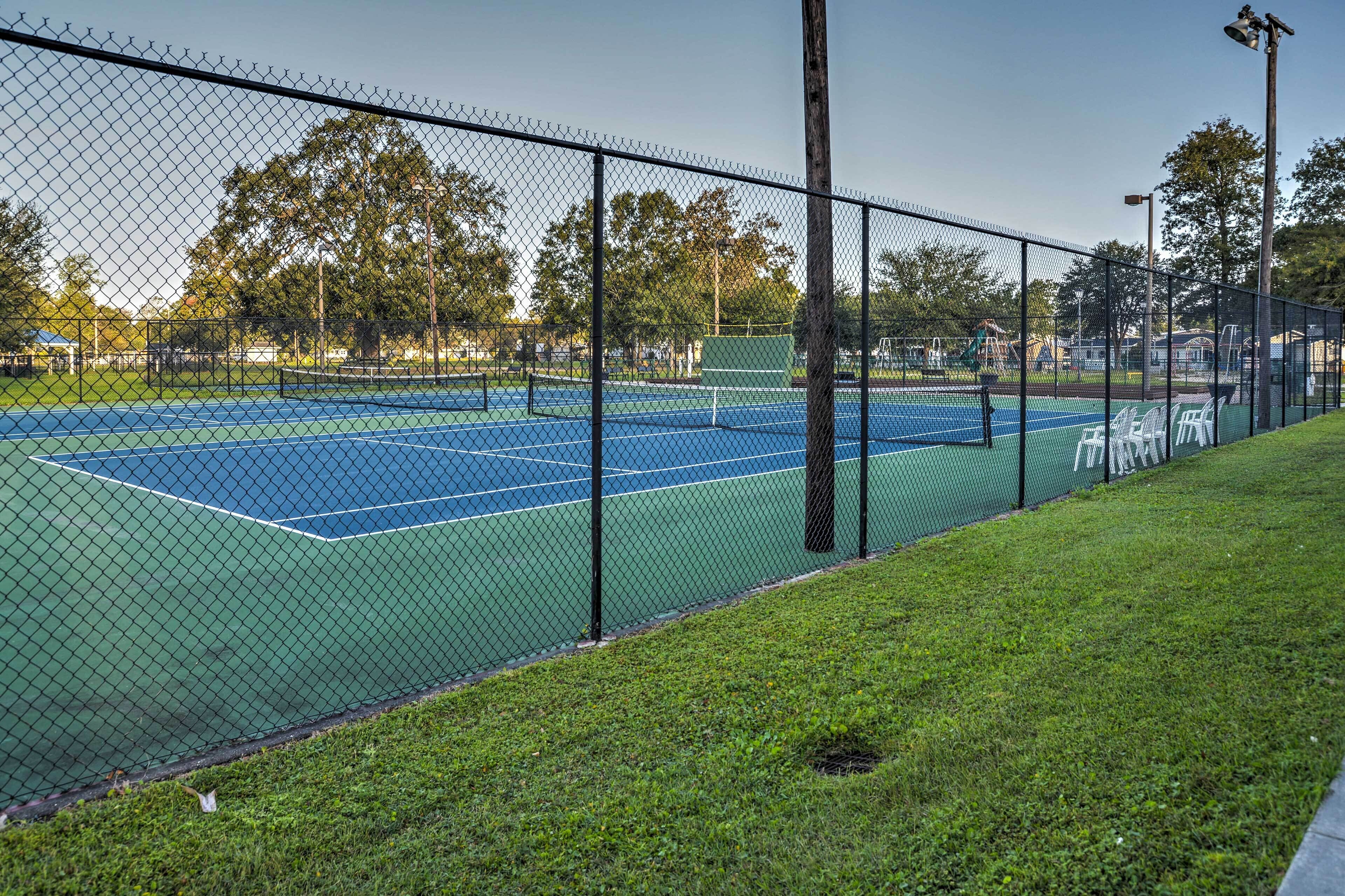 A tennis court with a chain-link fence, surrounded by grass and trees.