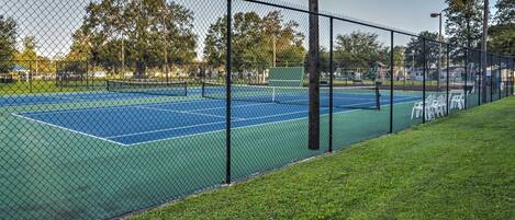 A tennis court with a chain-link fence, surrounded by grass and trees.