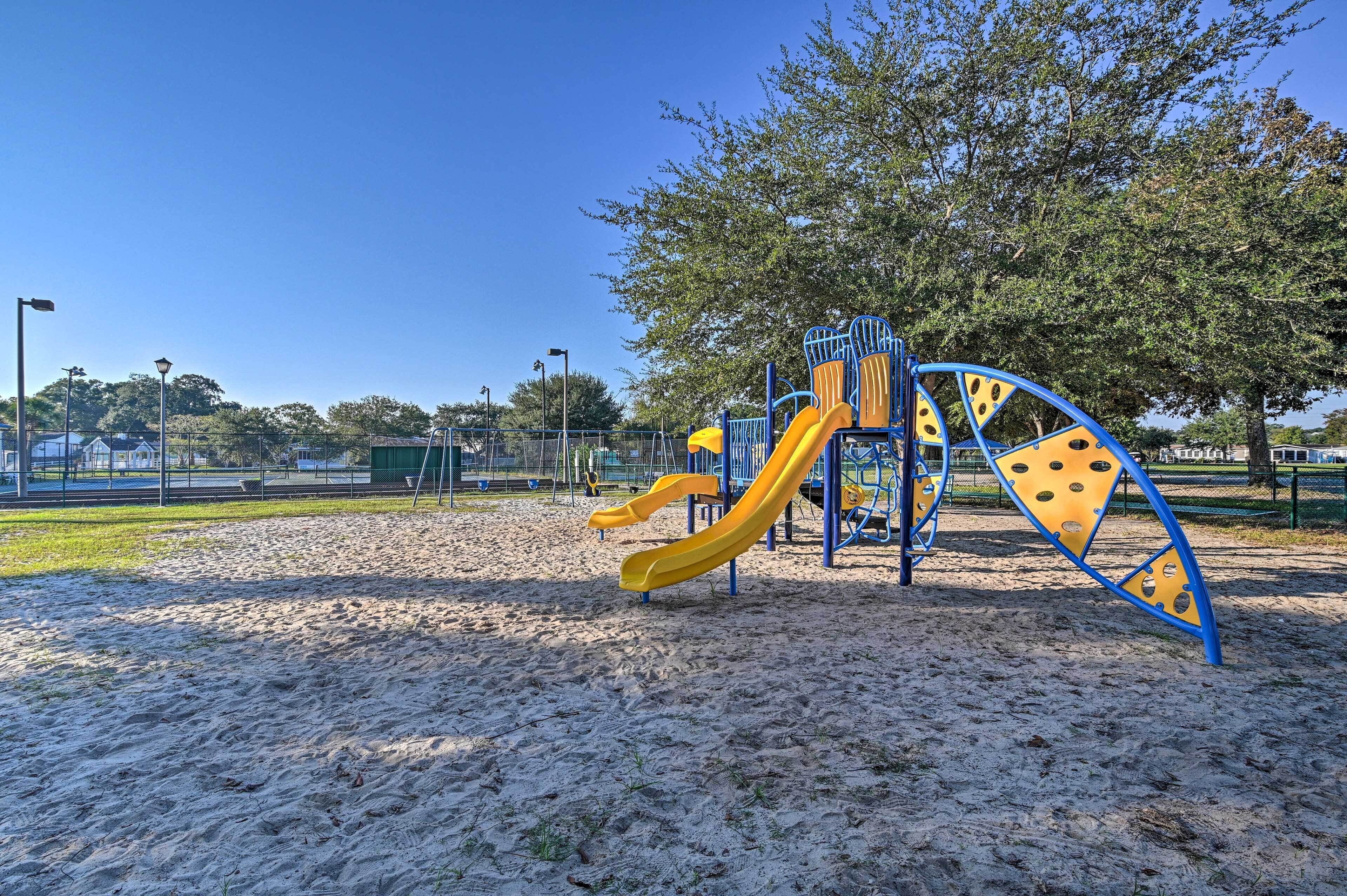 A playground with a yellow slide and blue climbing structures, set on sandy ground with trees in the background.