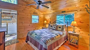 A cozy cabin bedroom with a bed, a ceiling fan, a nightstand with a lamp, a bookshelf, and a laundry area. - Cabin by Bold Creek: Book Now for Polar Express! (Bryson City)