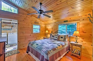 Ein gemütliches Schlafzimmer in einer Hütte mit Bett, Deckenventilator, Nachttisch mit Lampe, Bücherregal und einem Waschbereich.