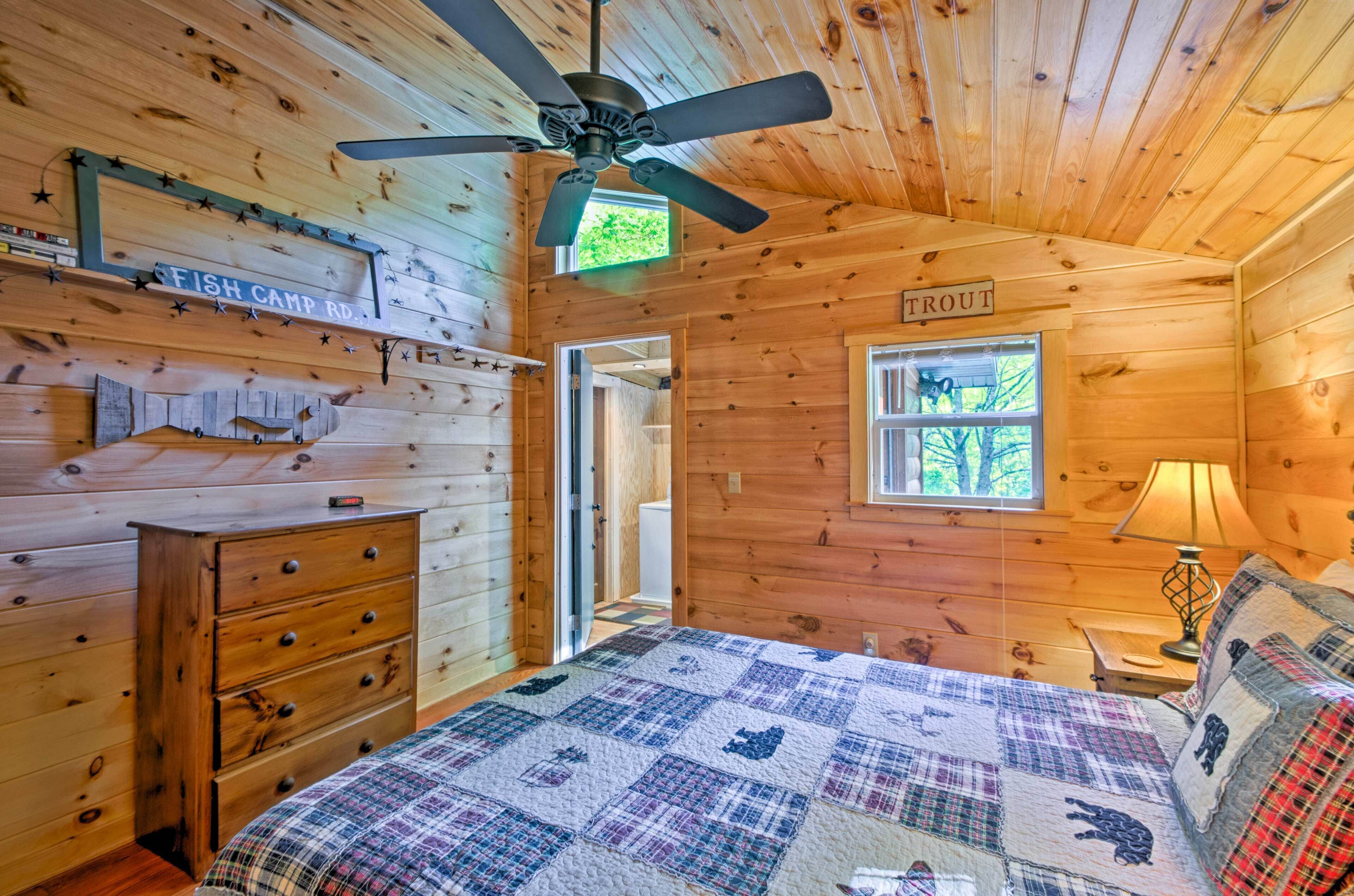 Interior de una cabaña con cama, cómoda, ventilador de techo y ventana con vista a árboles.