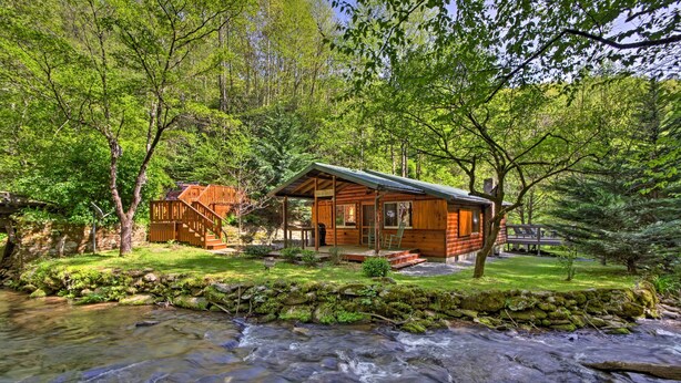 A wooden cabin with a porch and a stone wall, surrounded by a forest and a flowing stream. - Cabin by Bold Creek: Book Now for Polar Express! (Bryson City)