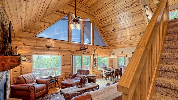 Un intérieur spacieux de chalet en bois, avec un plafond haut, de grandes fenêtres, un coin salon confortable et un escalier.