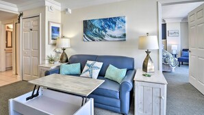 A living room with a blue sofa, a coffee table, and a painting of a wave. There are two lamps on side tables, and a door leading to another room.