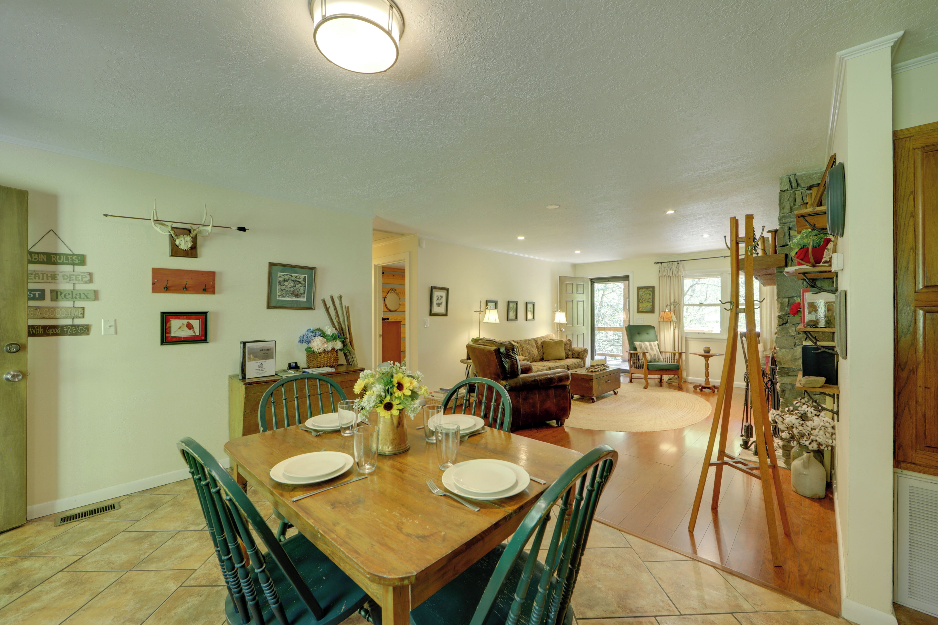 A dining area with a wooden table set for four, surrounded by green chairs. There is a vase with flowers on the table, and a painting on the wall.