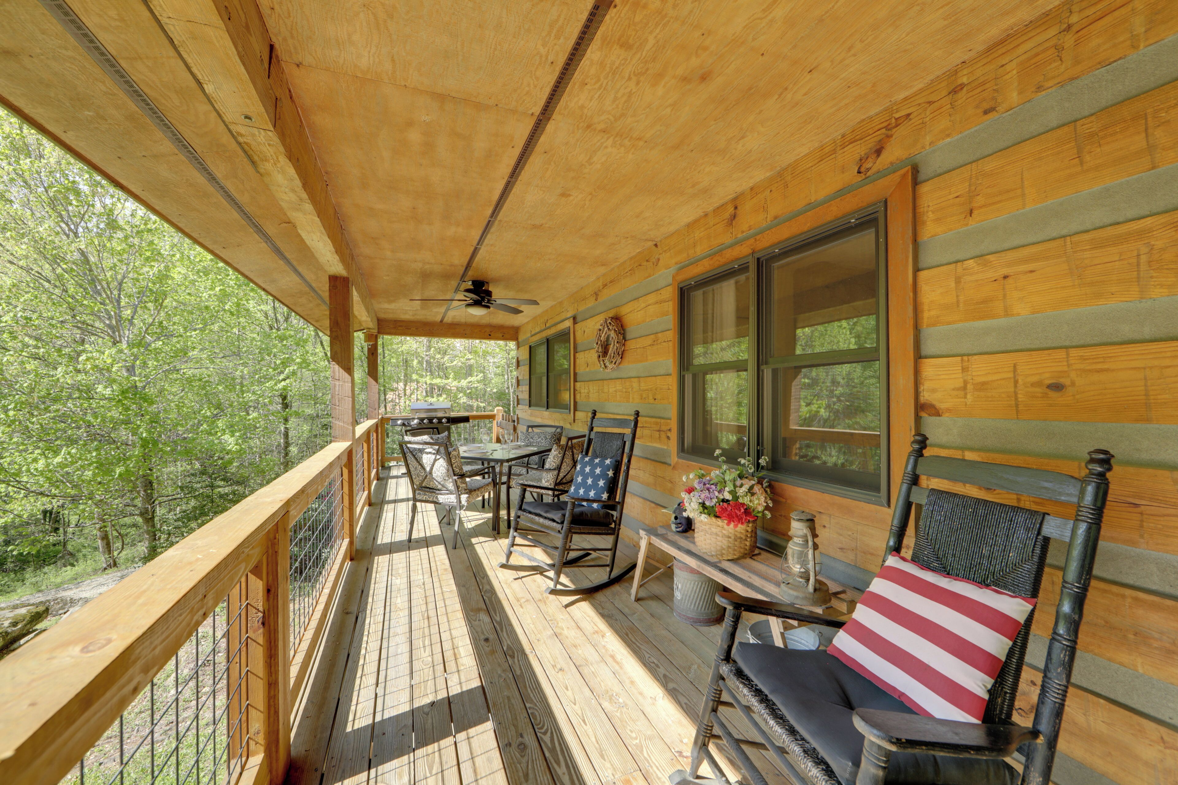 A wooden deck with a table and chairs, a wicker chair with a pillow, and a window with a view of trees.