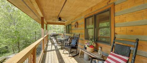 A wooden deck with a table and chairs, a wicker chair with a pillow, and a window with a view of trees.