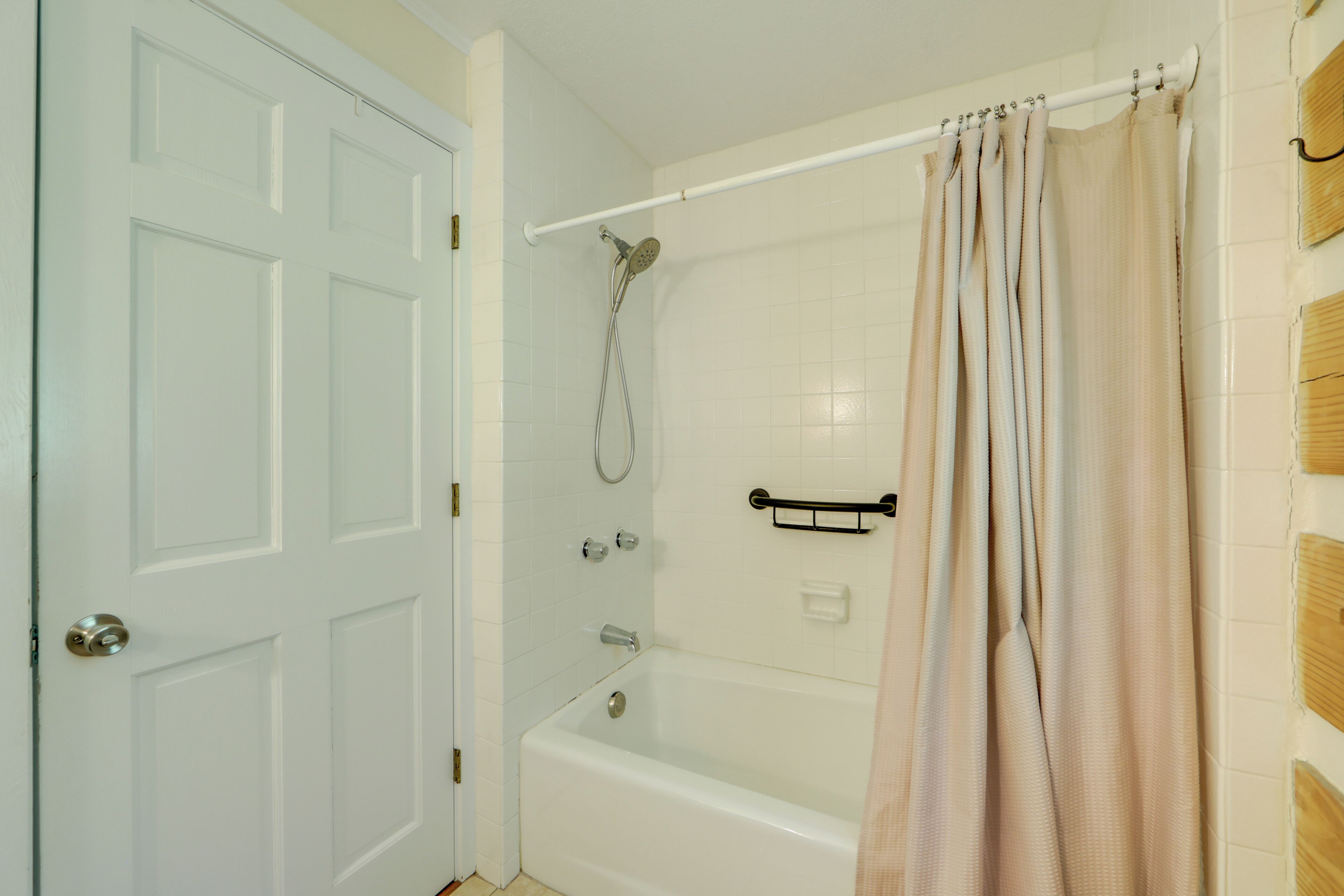 A bathroom with a white door, a bathtub, a showerhead, and a beige curtain.
