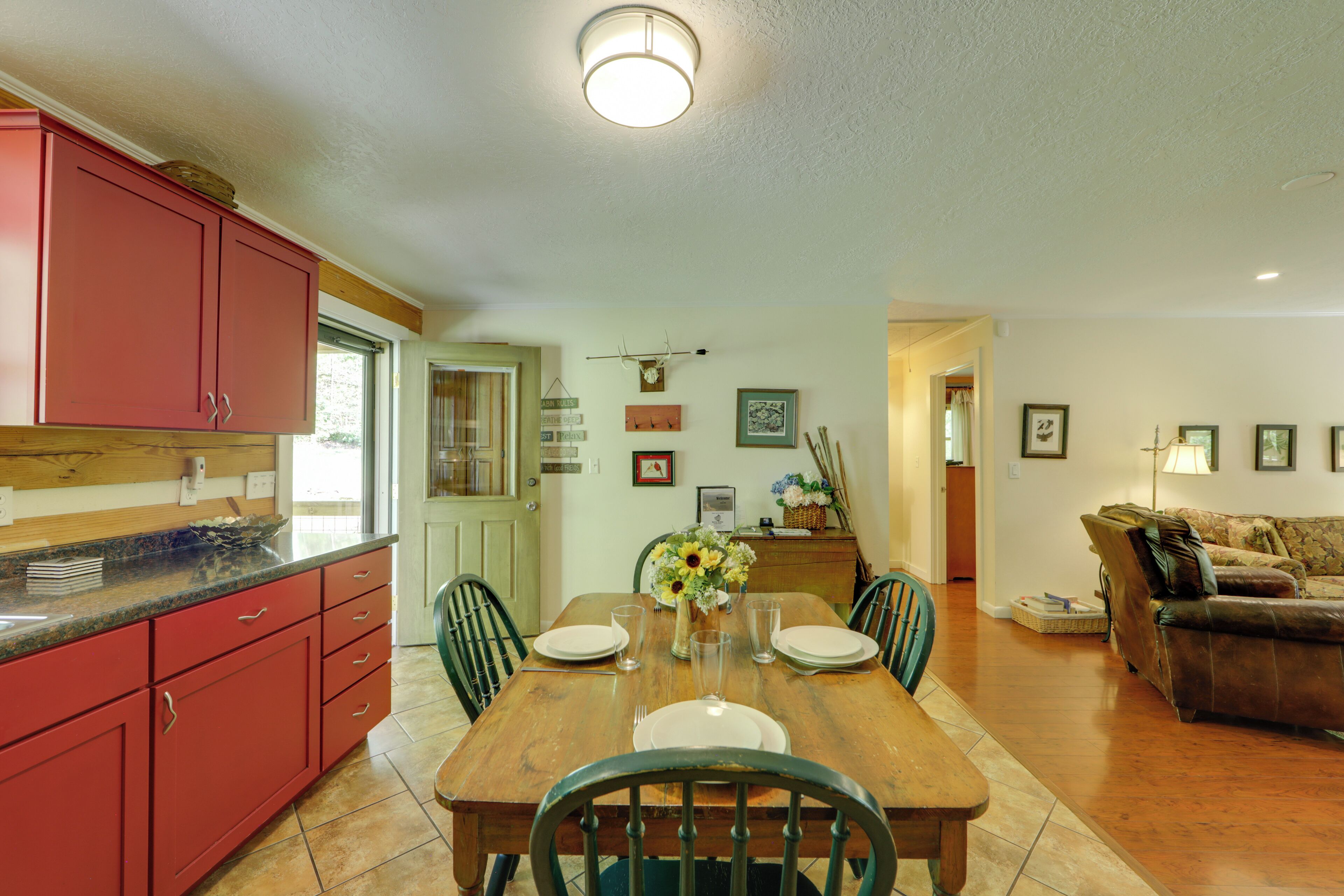 A dining area with a wooden table set for four, surrounded by chairs. A kitchen with red cabinets is visible to the left.