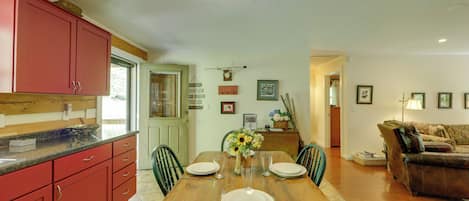 A dining area with a wooden table set for four, surrounded by chairs. A kitchen with red cabinets is visible to the left.