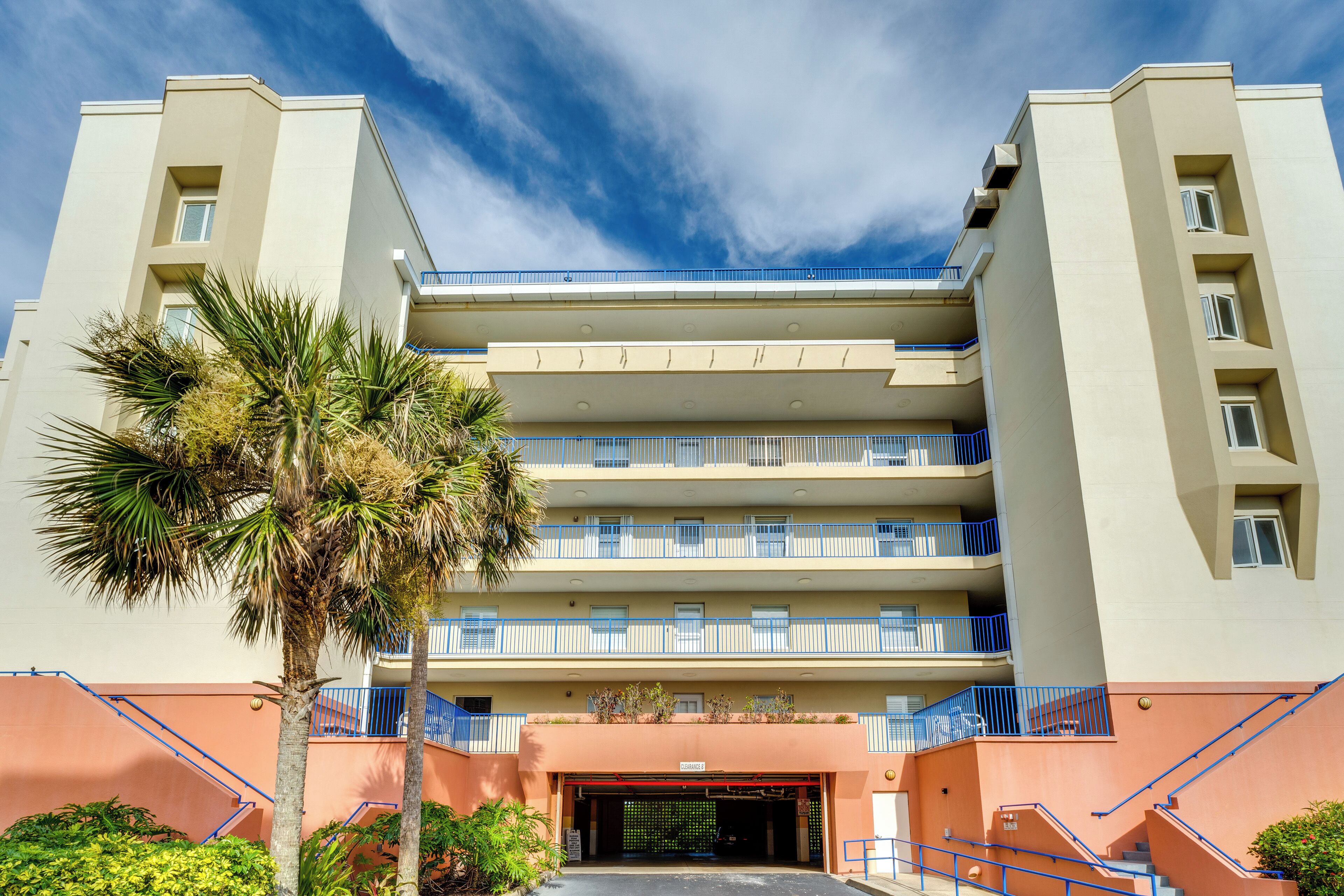 A multi-story hotel with balconies, a palm tree, and a clear sky.