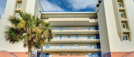 A multi-story hotel with balconies, a palm tree, and a clear sky.