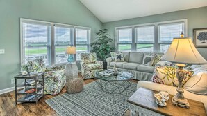 A living room with a view of a green field, a sofa, a chair, a coffee table, and a lamp. - Spacious Beach Cottage w/ Private Dock Access (Ocean Isle Beach)