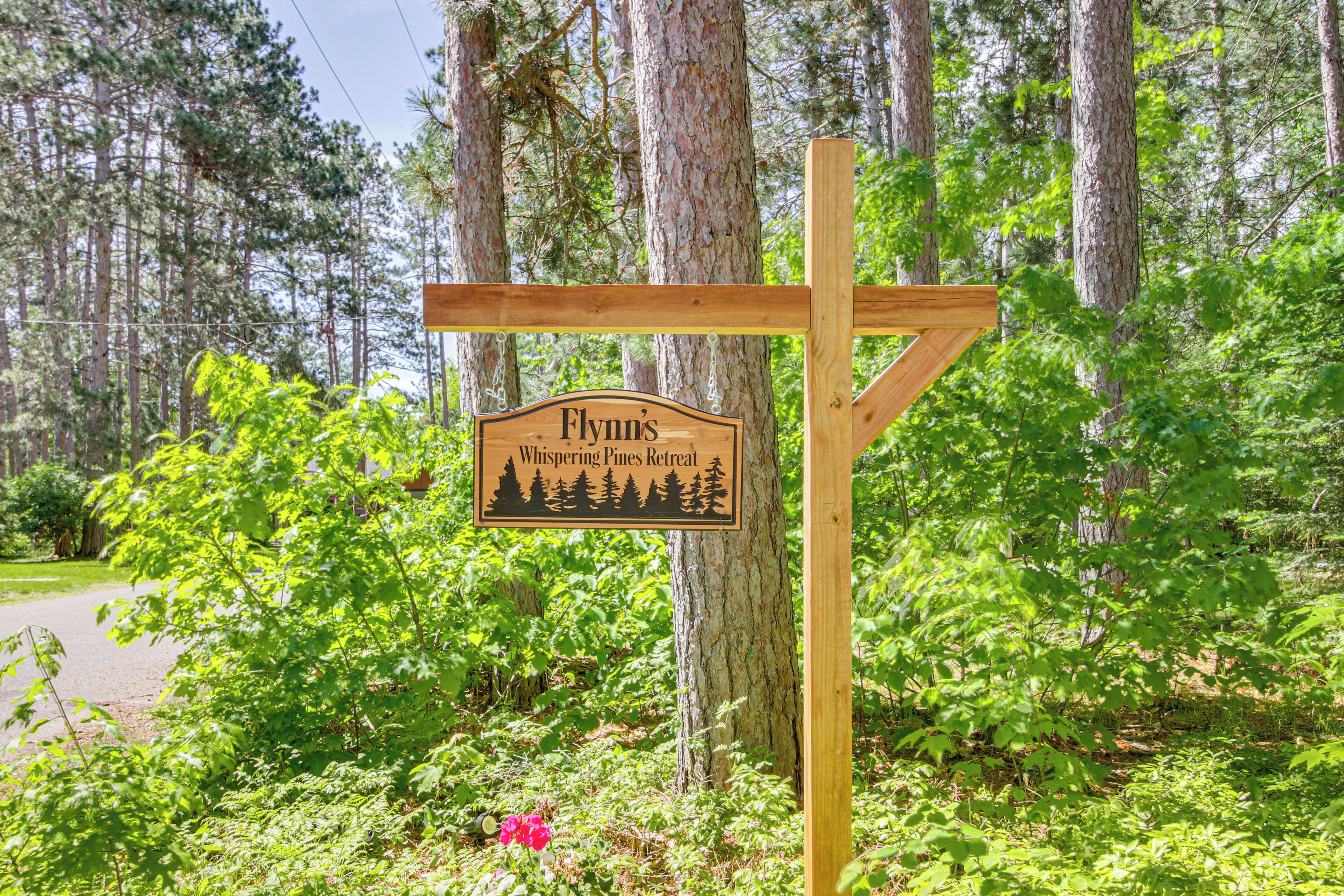 A wooden signpost with the text "Flynn's Whispering Pines Retreat" surrounded by lush greenery and trees.