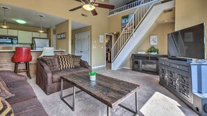 A spacious living room with a high ceiling fan, a staircase, a kitchen area, and a large TV.