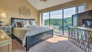 A bedroom with a large bed, a ceiling fan, a nightstand with a lamp, and a view of a lake through large windows.