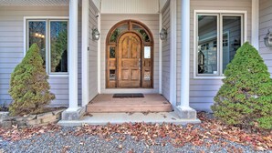 Une maison avec une porte d’entrée en bois, un porche aux colonnes blanches et deux petits arbres à feuilles persistantes.