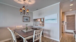A dining area with a dark wooden table, white chairs, a chandelier, and a kitchen visible through an open door. - Beachfront Condo w/ Unobstructed Ocean Views! (Oak Island)