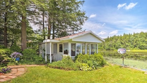 A small, single-story house with a porch, surrounded by lush greenery and a pond. - Cozy Cottage on Caroga Lake w/ Waterfront Fire Pit (Caroga Lake)