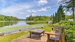 A lakeside outdoor seating area with a table and chairs, surrounded by lush greenery and a calm lake. - Cozy Cottage on Caroga Lake w/ Waterfront Fire Pit (Caroga Lake)