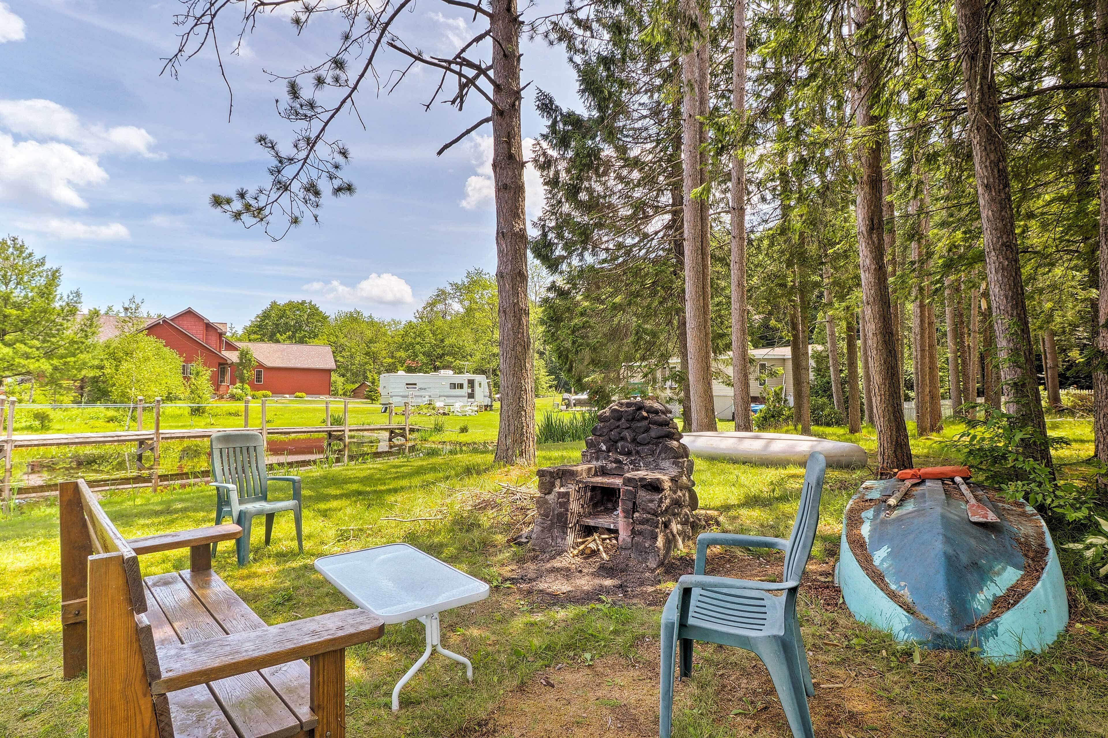 A rustic outdoor seating area with a fire pit, surrounded by tall trees and a red building in the background.
