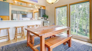 A spacious kitchen with wooden stools, a large wooden table, and a view of a forest through large windows. - Home w/ Hot Tub: 5 Mi to Angel Fire Bike Park (Angel Fire)