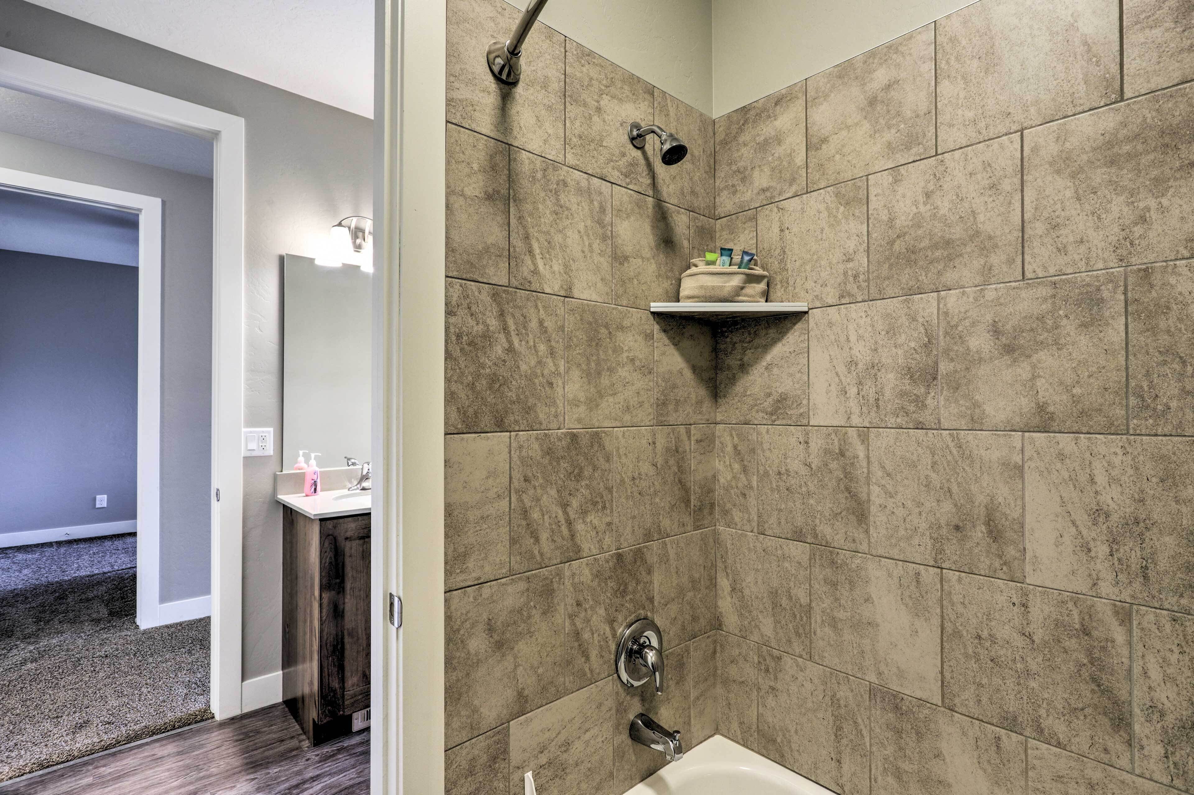 Un baño con azulejos color crema, una ducha, un lavamanos con artículos de aseo y un perchero para toallas.