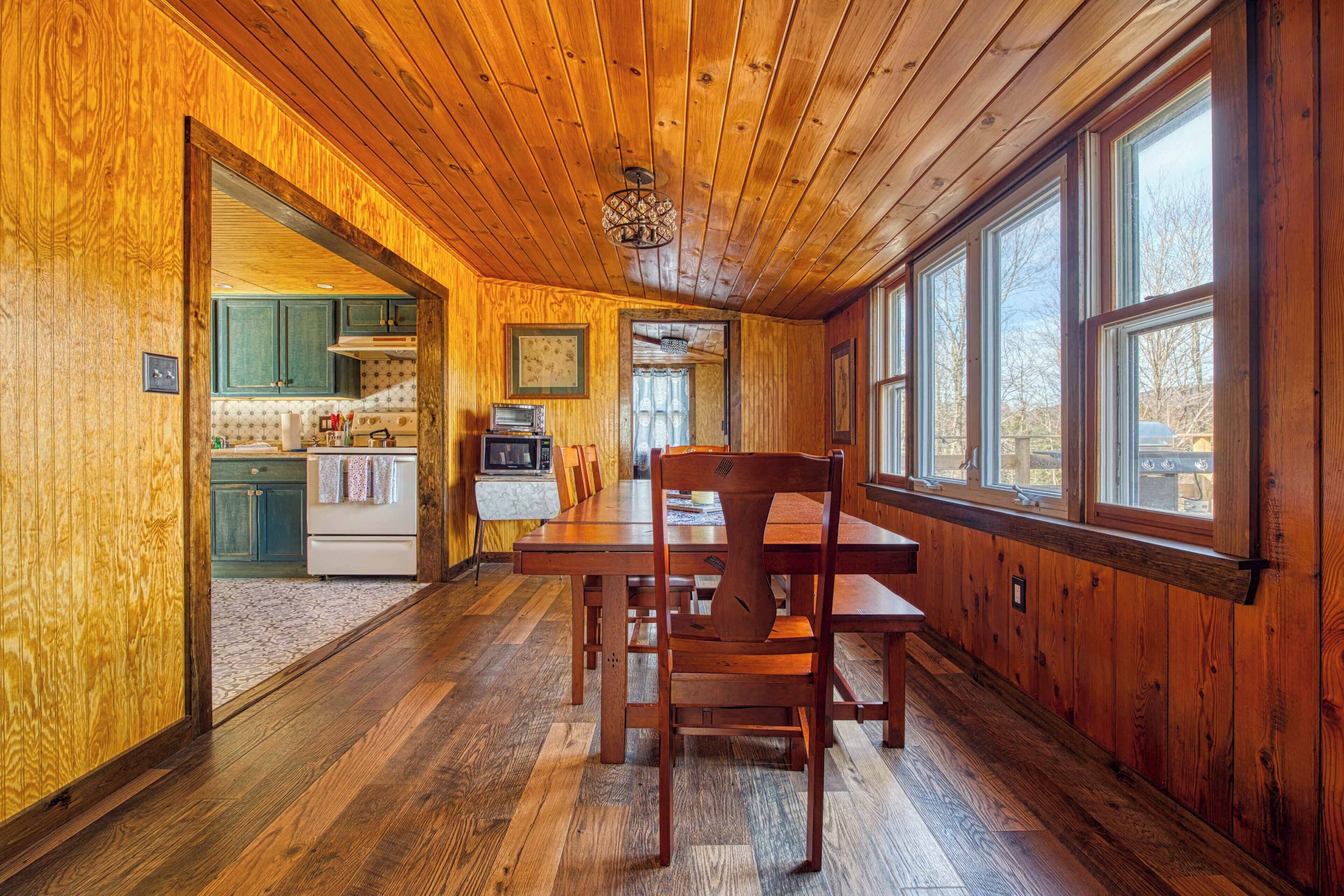 Interior de una cabaña de madera con comedor, cocina y grandes ventanales.