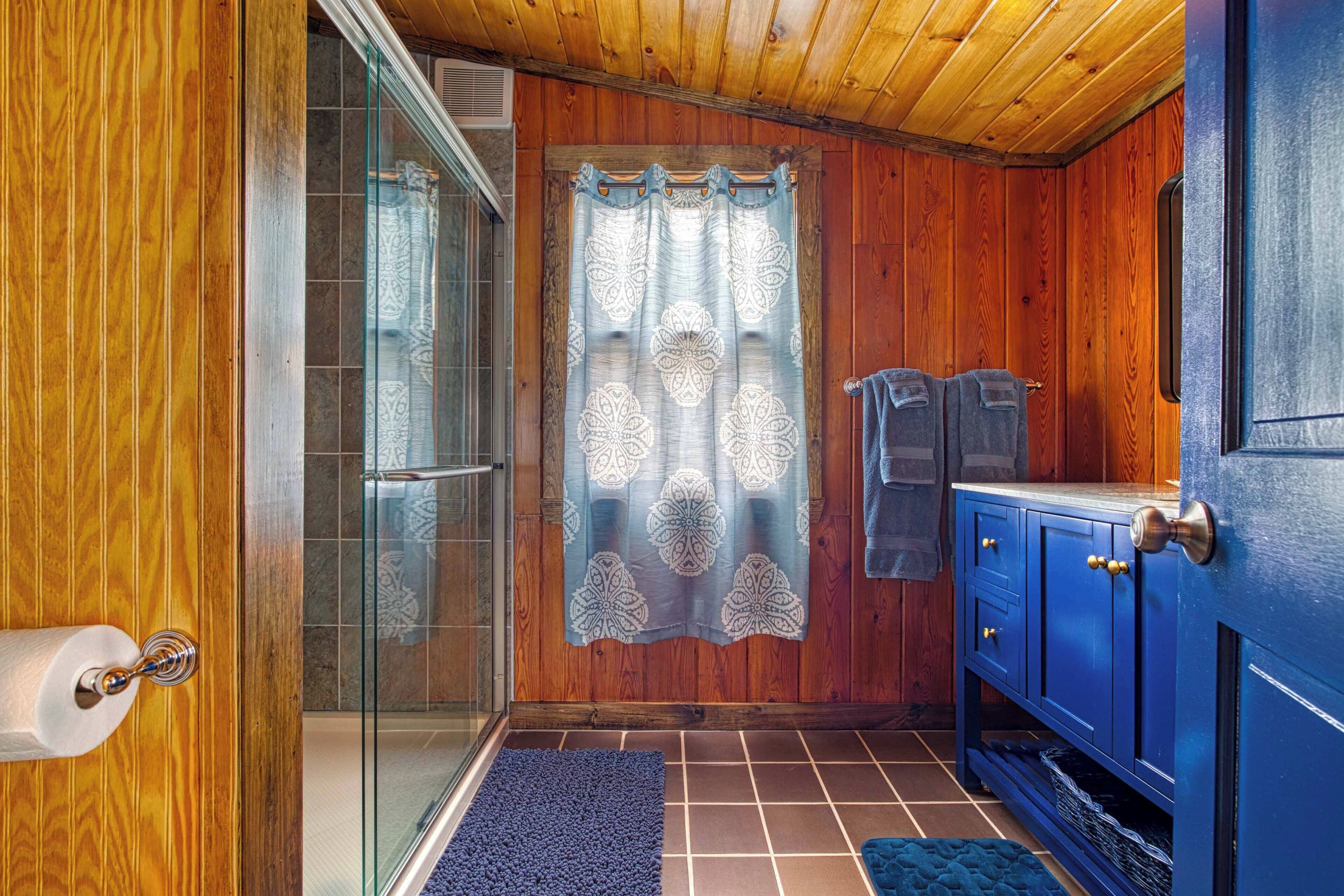 A wooden-paneled bathroom with a glass shower enclosure, a blue cabinet, and a patterned shower curtain.