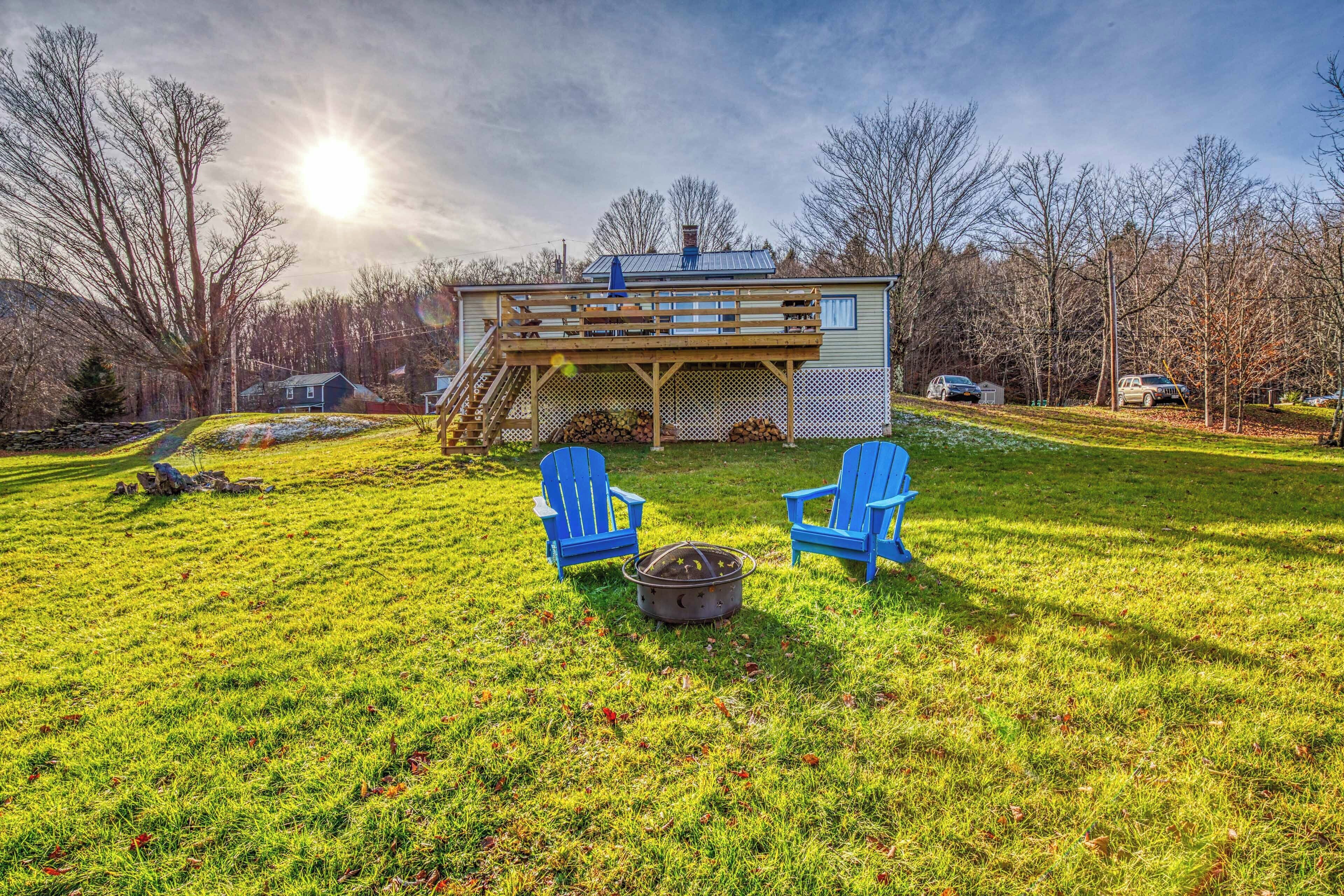 Ein Haus mit Terrasse, zwei blaue Adirondack-Stühle und einem Feuerkorb im Garten.