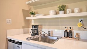 Eine Küche mit einer Spüle aus Edelstahl, einer Kaffeemaschine und einem Geschirrständer. Auf der Arbeitsfläche stehen zwei Flaschen Handseife.