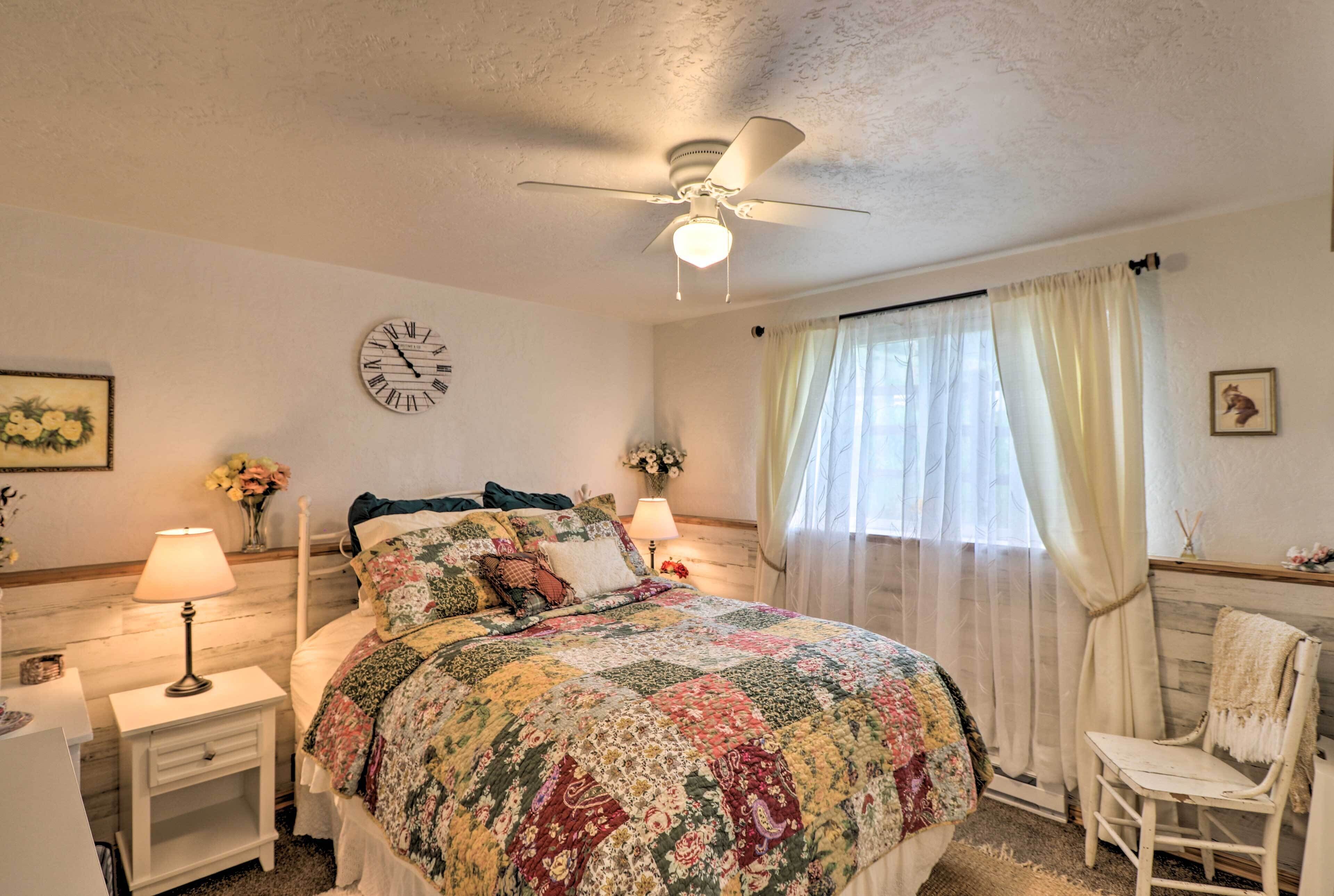 A bedroom with a bed, a nightstand with a lamp, a ceiling fan, a clock, a window with curtains, and a chair.