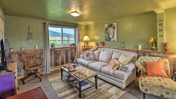 A cozy living room with a sofa, armchair, and a coffee table. There is a window with curtains, a lamp, and a painting on the wall. - Flathead Lake-view Studio: 38 Mi to Blacktail (Big Arm)