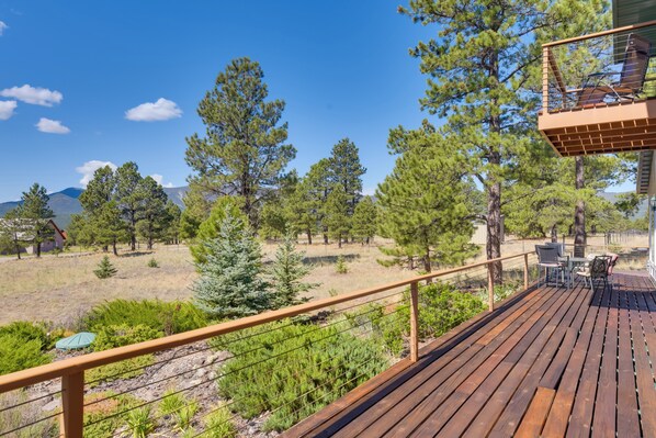 A wooden deck with outdoor furniture, overlooking a forested area with pine trees and a mountain in the distance.