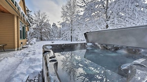 Un jacuzzi con tapa de vidrio transparente, rodeado de árboles cubiertos de nieve y un edificio de fachada color crema.