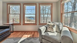 A room with a gray sofa, a wooden coffee table, and three large windows offering a view of trees and a lake. - Lake-view Retreat w/ Fire Pit in Mountain Home! (Mountain Home)