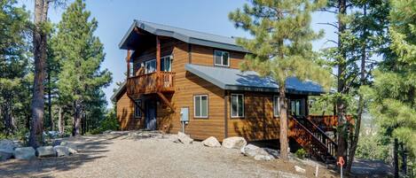 A two-story log cabin with a balcony, surrounded by pine trees.