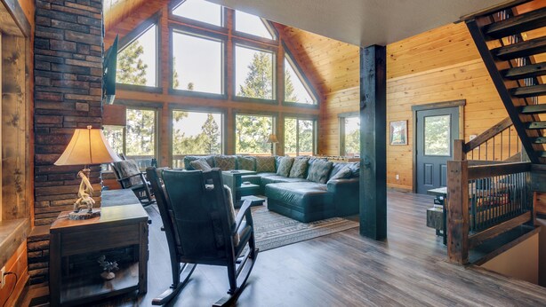 A spacious living room with a large sectional sofa, a rocking chair, and a fireplace. There are multiple windows providing natural light and a view of the outdoors. - 'longview Lodge' w/ Furnished Deck & Fire Pit! (Duck Creek Village)