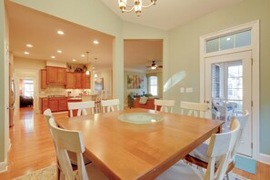 Un comedor amplio con una gran mesa de madera y sillas blancas, conectado a una cocina con armarios de madera y un ventilador de techo.